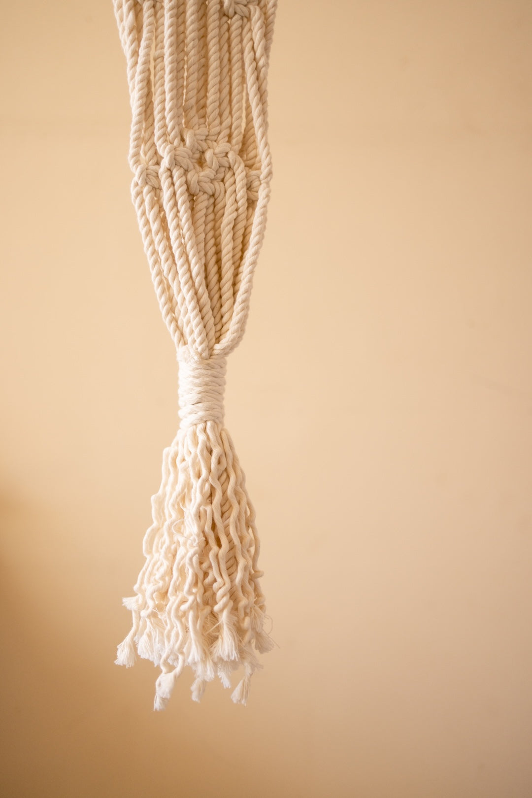 A cream macrame curtain rod hanger with intricate woven knots and a frayed tassel, shown hanging against a solid warm beige background.