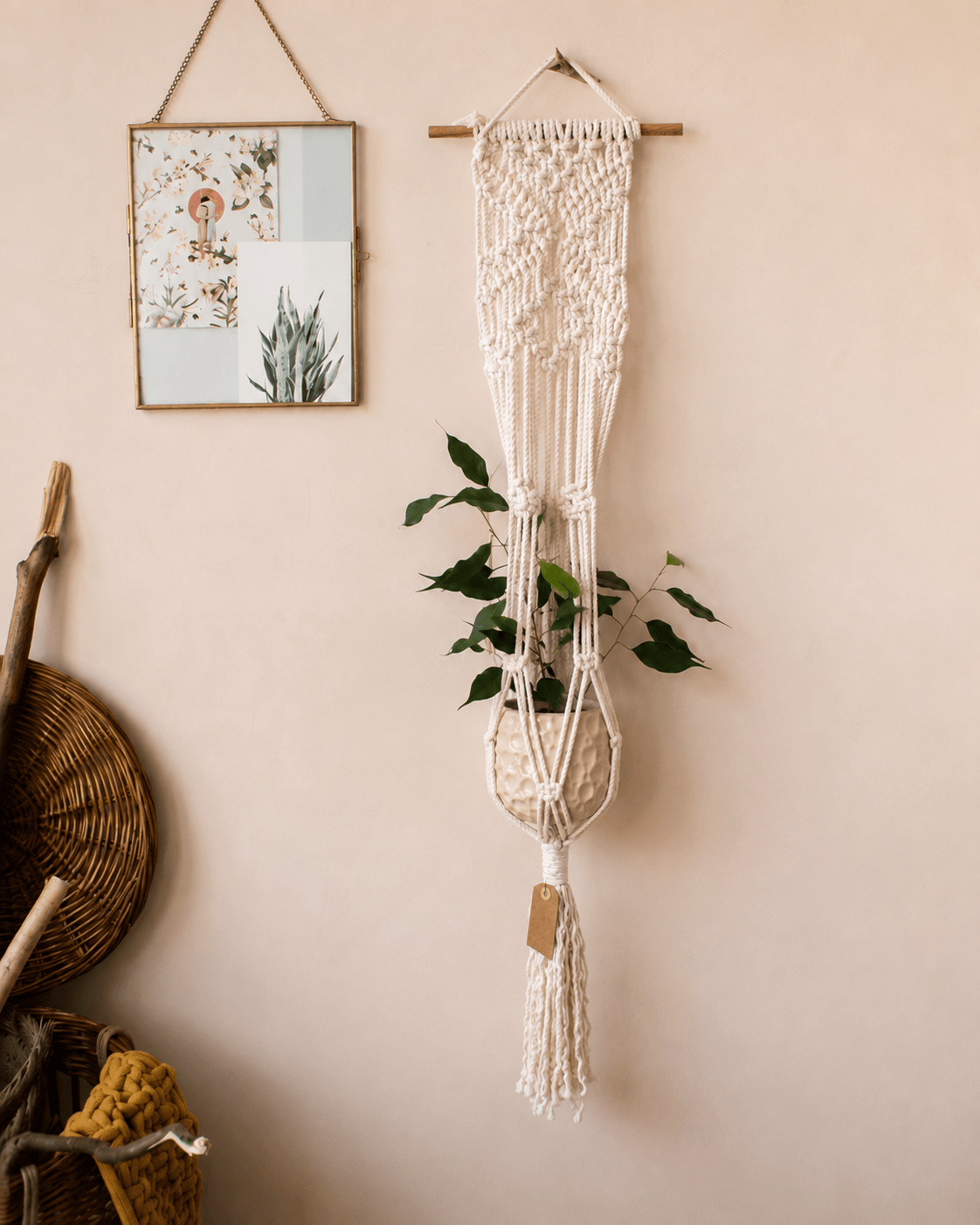 A cream macrame plant hanger with a wooden rod and tassel, holding a small potted green plant, hanging on a beige wall next to a framed print.