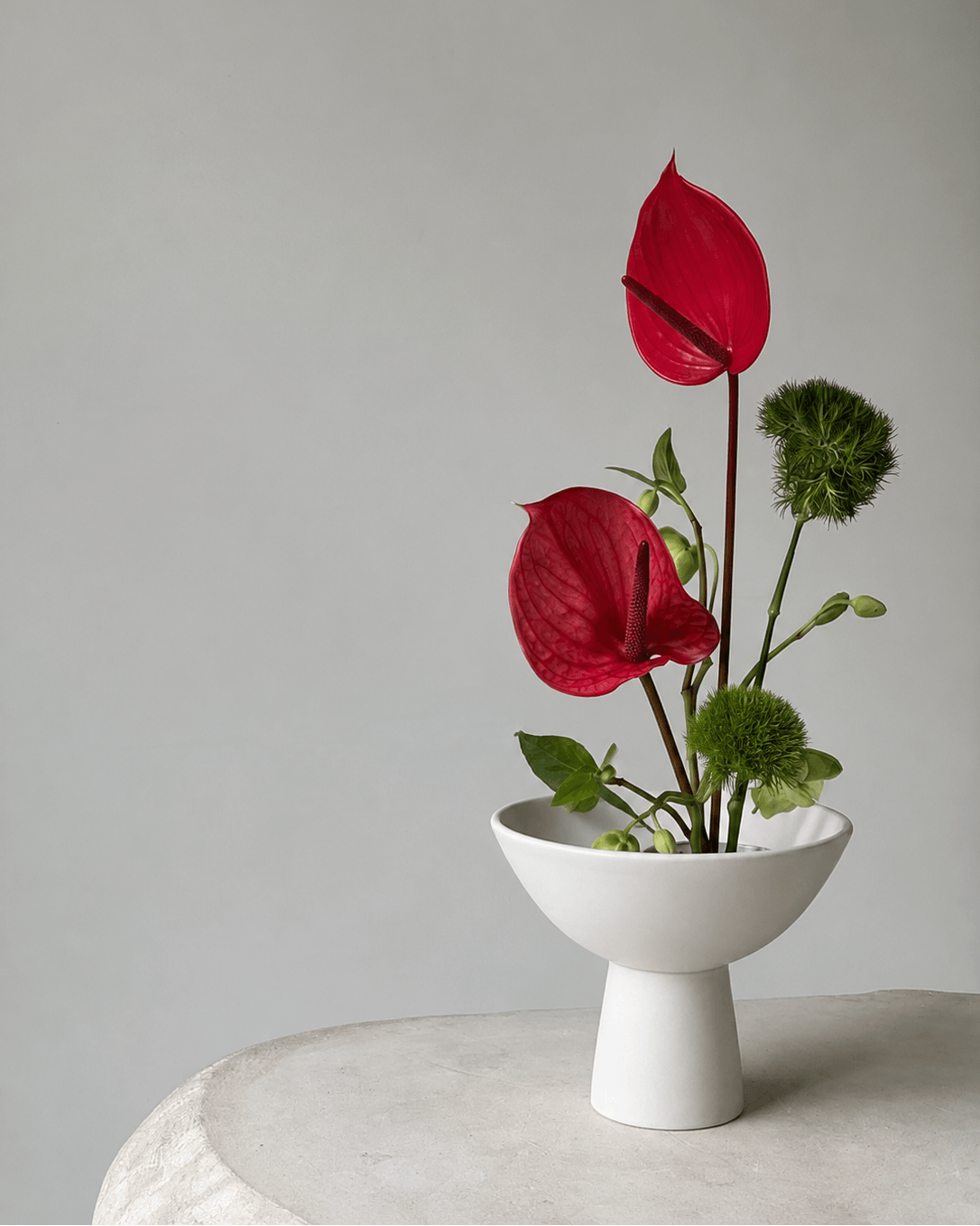 A minimalist floral arrangement featuring two bright red anthuriums and green textured filler stems in a white ceramic bowl-shaped vase, set on a light stone surface against a soft gray background.