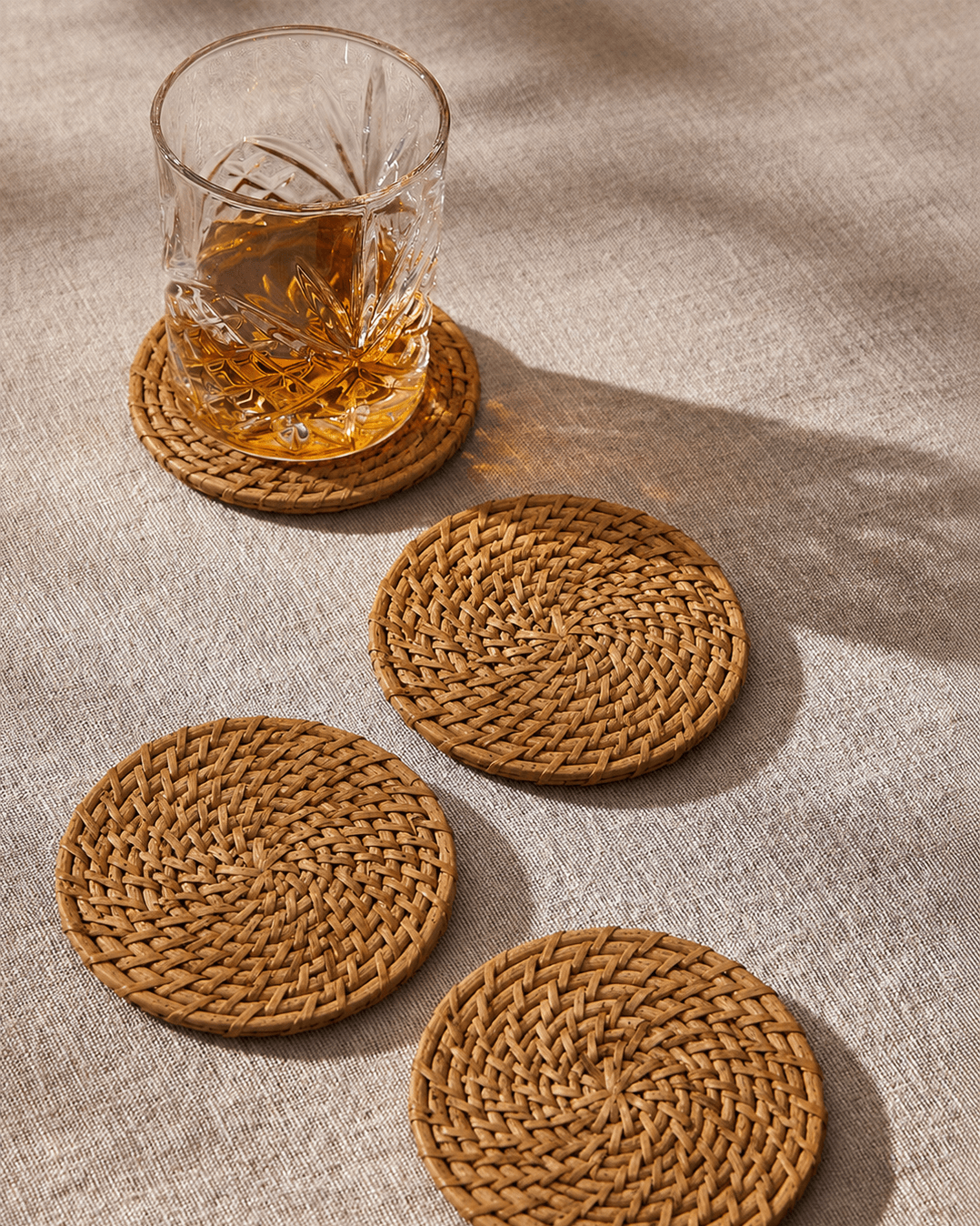 Three round woven natural fiber coasters in a warm tan-brown color, arranged on a textured beige fabric surface. One coaster holds a clear cut-glass tumbler filled with amber liquid, and the scene is lit with warm directional sunlight casting soft shadows.