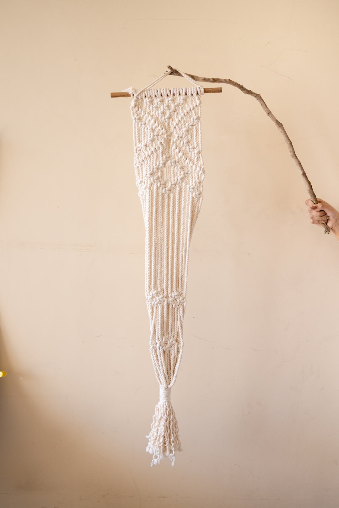 An off-white macrame wall hanging suspended from a wooden dowel, shown against a plain beige wall with a hand holding a branch on the right.