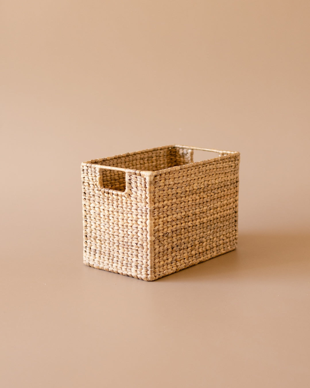 A small woven rectangular storage basket with cutout handles, made from water hyacinth, a natural straw or seagrass, set against a solid warm beige background.